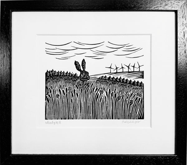 Framed lino print in black ink of hare in long grass and wind turbines on distant hill