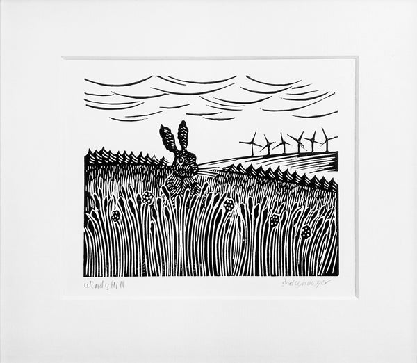 Mounted lino print in black ink of hare sitting in long grass and wind turbines on distant hill